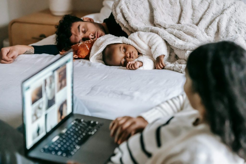 A cozy scene of family bonding with a sleeping baby and laptop in the bedroom.