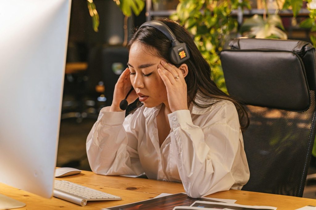 Asian woman in office feeling overworked, wearing a headset, sitting at a computer desk with a tired expression.